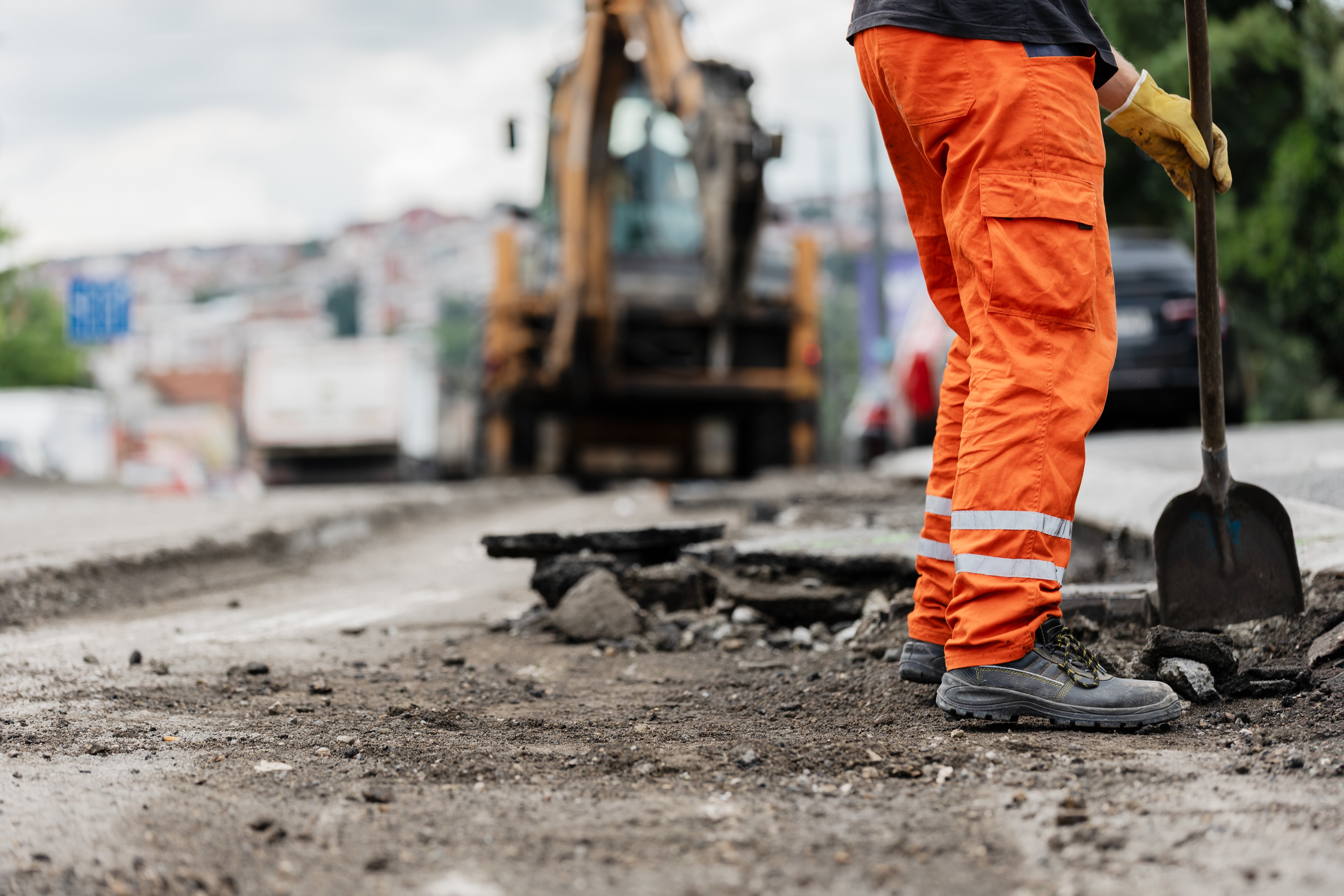 A construction worker in bright orange pants uses a shovel to repair the road surface, with heavy machinery in operation nearby under overcast skies and urban setting.