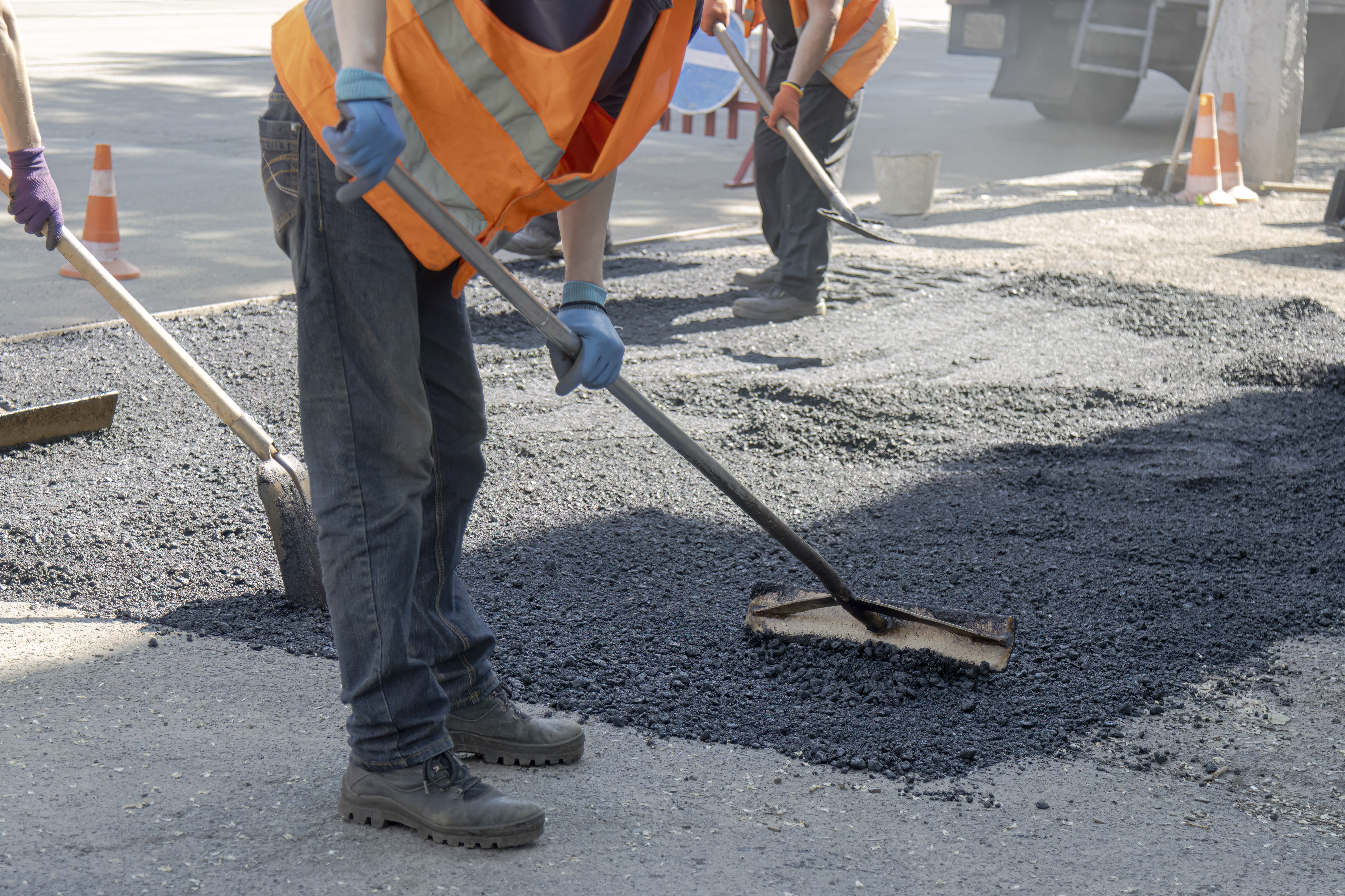 A crew of road maintenance laborers working on a street renovation project. In the foreground, a construction worker wearing a high-visibility orange vest and protective gloves bends down to smooth and flatten hot black bitumen using a specialized asphalt scraper tool. The background features other workers with shovels, a truck, and traffic cones, highlighting urban infrastructure development, civil engineering, manual labor, and municipal teamwork
